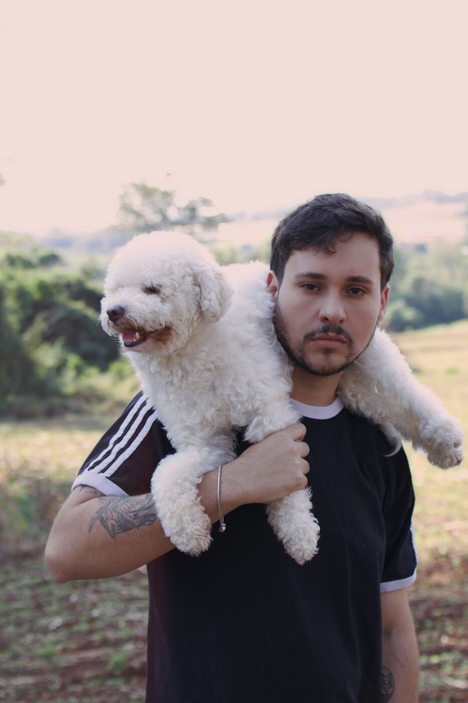 Homem com um poodle branco nos ombros, num campo aberto no fim da tarde